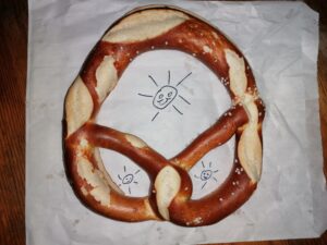 Laugenbrezel auf einer Papiertüte, auf die drei Sonnen gemalt sind, die durch die Öffnungen der Brezel hindurchscheinen