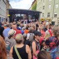 Viele Personen stehen vor einer Bühne auf dem Rio-Reiser-Platz, auf der »Ton Steine Scherben« spielen. Davor ein Transparent »Keine Macht für niemand«
