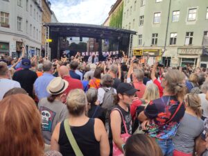 Viele Personen stehen vor einer Bühne auf dem Rio-Reiser-Platz, auf der »Ton Steine Scherben« spielen. Davor ein Transparent »Keine Macht für niemand«