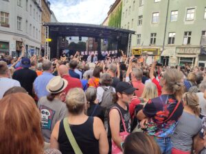 Viele Personen stehen vor einer Bühne auf dem Rio-Reiser-Platz, auf der »Ton Steine Scherben« spielen. Davor ein Transparent »Keine Macht für niemand«