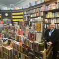 Raumfüllendes volles Bücherregal, davor ein mit Bücher überquellender Verkaufstresen, lächelnd daneben Buchhändler Harald Kirchner