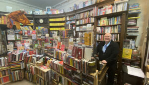 Raumfüllendes volles Bücherregal, davor ein mit Bücher überquellender Verkaufstresen, lächelnd daneben Buchhändler Harald Kirchner