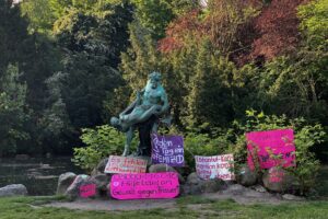 Ernst Herters Bronzeskulptur »Ein seltener Fang« im Viktoriapark: Ein Fischer ringt mit einer Meerjungfrau, die er aus dem Wasser gezogen hat. An der Skulptur lehnen diverse Pappschilder: »Jeden 3. Tag ein Femizid«, »Es fehlen 15.000 Frauen*hausplätzer«, »08000116016 Hilfetelefon Gewalt gegen Frauen*«, »Instanbul-Konvention konsequent umsetzen«, »Schutzräume schaffen und erhalten«