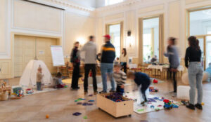 Erwachsene und Kinder im Saal des Nachbarschaftshauses mit Spielmaterial auf dem Boden und einem kleinen Spielzelt.