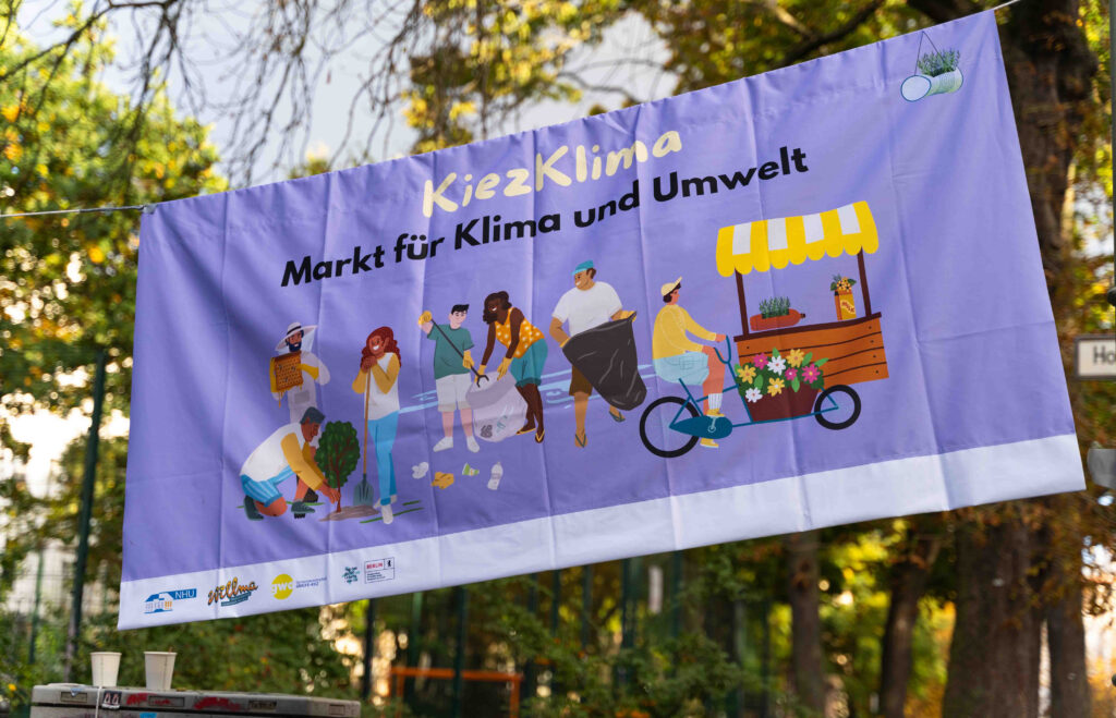 Zwischen Bäumen aufgehängtes Banner mit der Aufschrift „Markt für Klima und Umwelt“ und Illustrationen nachhaltiger Alltagsaktivitäten.