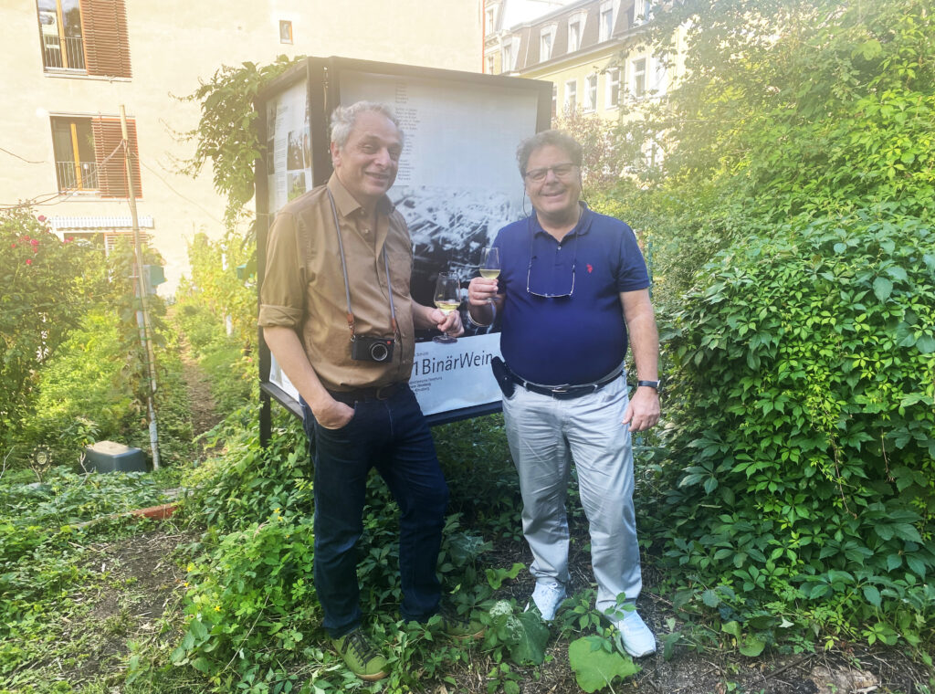 Martin G. Schicht und Dr. Anagnostis Paraskevopoulos stehen zwischen Weinreben und halten ein Schild des Projekts Z1BinärWein.